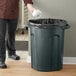 A man in a chef's uniform throwing a white object into a green Toter round trash can.