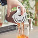 A person using a Franmara stainless steel cocktail strainer to pour a drink into a glass.