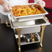 A person holding a Vollrath water pan of pasta and sausages in front of a classic brass chafer on a table in an outdoor catering setup.