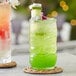 A green Acopa Tiki glass filled with a green cocktail with a tropical fruit garnish on a woven mat.