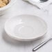 A white CAC scalloped edge demitasse saucer on a white surface with a bowl and spoon.