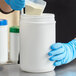 A hand in a blue glove pouring white powder into a white HDPE plastic canister.