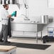 A man in a black apron and gloves standing in front of a Regency stainless steel 3 compartment sink on a counter in a professional kitchen.