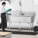A woman in a black apron using a Regency stainless steel three compartment sink in a professional kitchen.