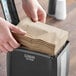 A person putting a Dixie Ultra Brown interfold paper napkin into a dispenser on a counter in a professional kitchen.