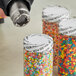 A group of jars with white 1" perforated shrink bands with black tamper-evident text over jars of colorful candy sprinkles.