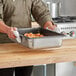 A person holding a Vollrath stainless steel steam table pan filled with food.