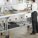 A man and woman using a ServIt electric steam table with a sneeze guard in a school kitchen.