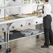 A man and woman standing behind a ServIt open well steam table with a glass sneeze guard.