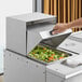 A hand using a Choice stainless steel roll top cover to close a tray of food with broccoli and carrots.