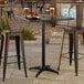 A black powder-coated aluminum bar height outdoor table with a square top and umbrella hole, shown with two metal bar stools.