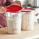 A woman pouring white liquid into a Vigor clear polycarbonate food storage container with a red lid.