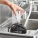 A hand using a black AvaMix rinser to pour water into a blender jar on a counter.