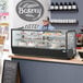 An Avantco black refrigerated countertop bakery display case on a counter with cakes and desserts, with a man behind the counter.