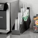 A black ServSense countertop cup, lid, and straw organizer holding clear plastic cups.