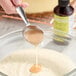 A spoonful of pineapple bakery emulsion being poured into a bowl of batter, with a bottle of LorAnn Oils Pineapple Bakery Emulsion in the background.
