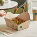 A hand holding a New Roots Kraft PLA-Lined take-out box of food on a table.