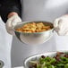 A person in white gloves holding a Vollrath stainless steel mixing bowl of food.