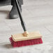 A person using a Lavex 10" bi-level floor scrub brush with a handle to clean a tile floor.