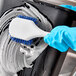 A person in blue gloves using a white and blue Lavex pot scrub brush to clean a black container.