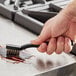 A hand using a Lavex toothbrush style grout brush with brass bristles to clean a metal surface.