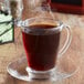 A glass cup of Ellis Mezzaroma Breakfast Blend coffee with steam coming out of it on a saucer.