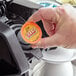 A hand holding a round orange Ellis Red Eye High Caffeine Coffee single serve cup with a grey lid.
