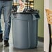 A person standing next to a Rubbermaid grey trash can with a black bag on top.