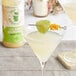 A bottle of Top Hat Provisions Classic Margarita Mix on a table with a glass of white liquid and a lime on top.