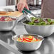 A person using tongs to mix vegetables in a Choice stainless steel mixing bowl.