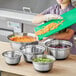 A woman using a green cutting board to cut vegetables into a silver mixing bowl.