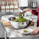 A woman pouring salad greens into a Choice stainless steel mixing bowl on a kitchen counter.