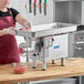 A woman in a red apron using a ProCut stainless steel electric meat grinder on a counter in a commercial kitchen.