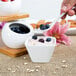 A hand holding a white American Metalcraft square porcelain sauce cup with a spoon full of blueberries over a bowl of yogurt.