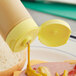 A close-up of a ham and cheese sandwich with mustard being poured on it using a yellow squeeze bottle lid.