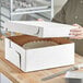 A person holding a white corrugated bakery box with a white cake inside.