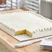 A large rectangular sheet cake with white frosting on a white corrugated cake board.