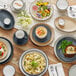 A table with Acopa Embers Harvest Tan stoneware bowls and plates of food, including a bowl of pasta with vegetables and cheese.