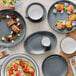 A grey table with Acopa midnight blue matte coupe stoneware platters and bowls of food.