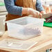 A person wearing gloves cutting vegetables in a Vigor translucent plastic food pan on a white counter.