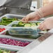 A person in gloves holding a Vigor translucent polypropylene food container filled with cucumbers.