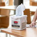 A hand cleaning a table with a white Rubbermaid container holding white disposable microfiber cloths.