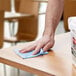 A hand wiping a table with a blue Rubbermaid disposable microfiber cloth.