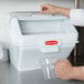 A person using a clear glass to pour ingredients into a white Rubbermaid ingredient bin.