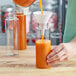 A hand pouring orange liquid into a clear cylinder bottle.