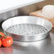 An American Metalcraft tin-plated steel pizza pan with holes on a wooden surface next to tomatoes and a cutting board.