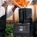 A man in a white shirt pours carrots into a Sammic batch bowl food processor on a counter in a professional kitchen.