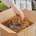 A hand holding a box of Spring-Fill Kraft Crinkle Cut Paper Shred over a cardboard box.