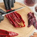 A person wearing black gloves is slicing open a dried guajillo chile pepper on a wooden cutting board.