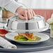 A hand using a Choice stainless steel plate cover to lift a plate of food.